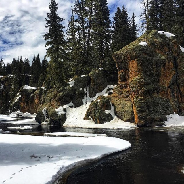 A narrow stream meanders through a snowy valley with rock spires and evergreen trees.