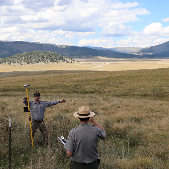 A park ranger holds a GPS device near a road while another park ranger takes a photo of the site.