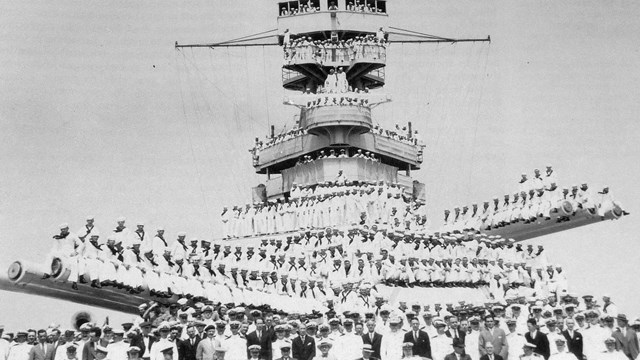 A black and white battleship sits with people around it.