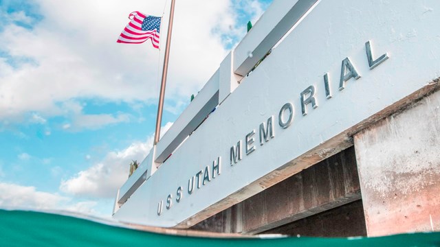A blue wave in front of the white memorial that reads USS Utah Memorial and an American flag