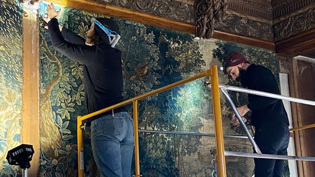 Two conservators on scaffolding removing large tapestries from a paneled wall.