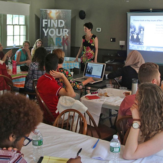 A group of professionals is seated listening to a teacher talk