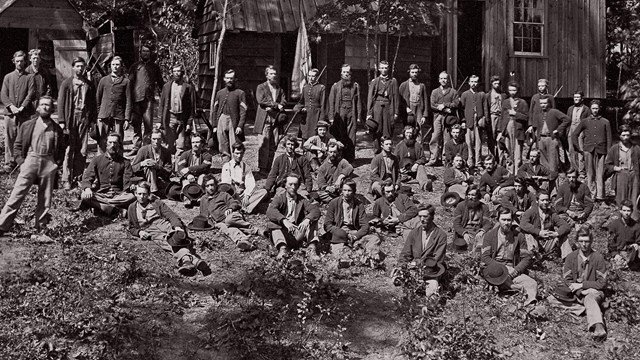 Federal soldiers standing in front of a house with pine trees in the background.