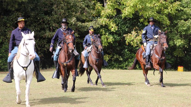Four men dressed in USCT uniforms ride horses 