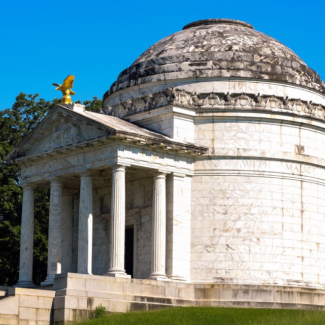 A large white marble structure with a dome roof sits atop a small grassy hill.