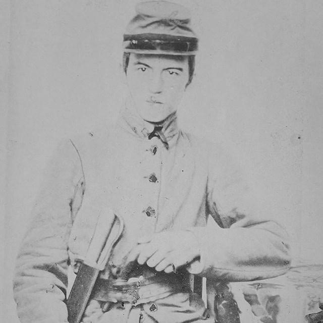 A Confederate soldier with gray cap and uniform with a bowie knife in their belt.