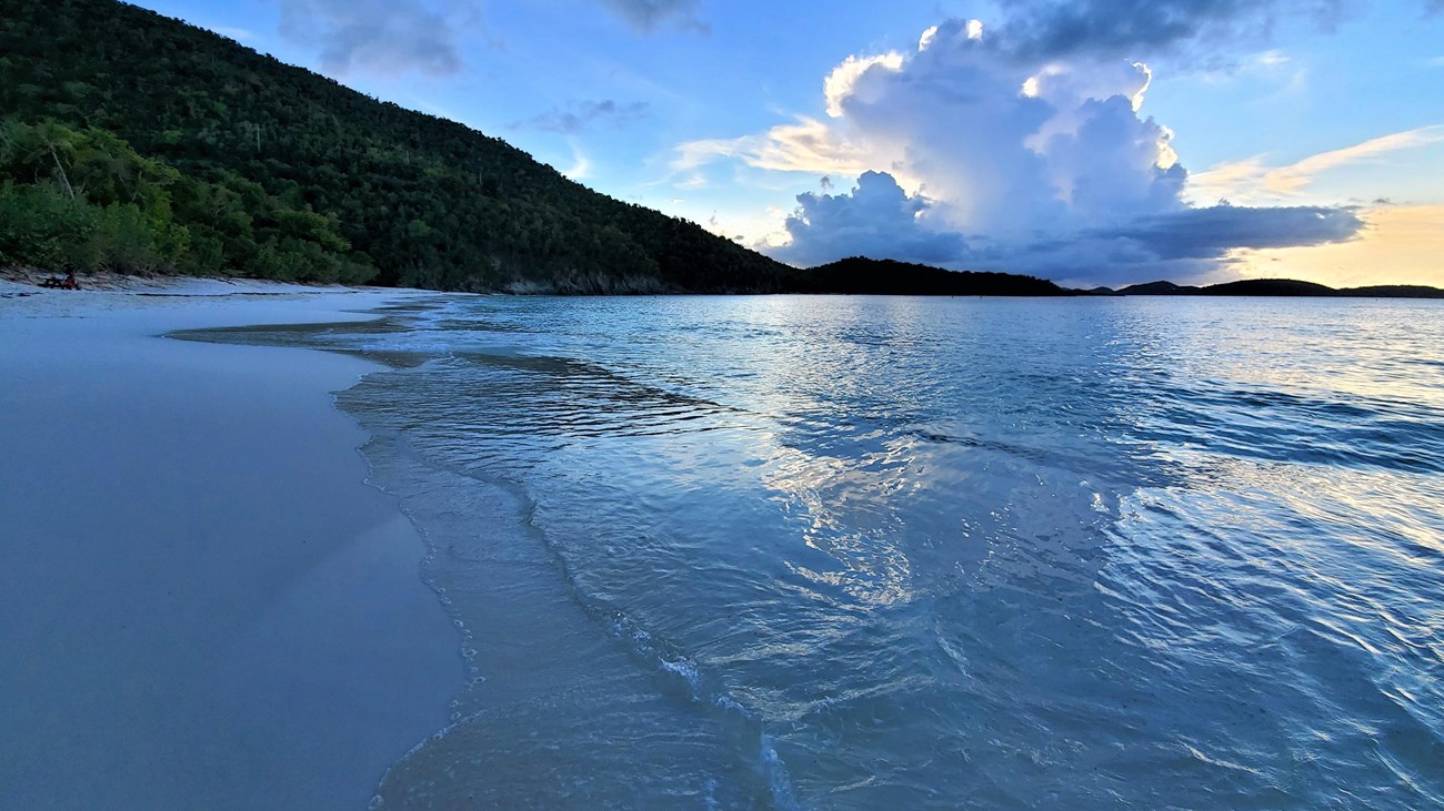 A white-sand beach with a small wave coming to shore. A cloud tints the low sun at base of hill..