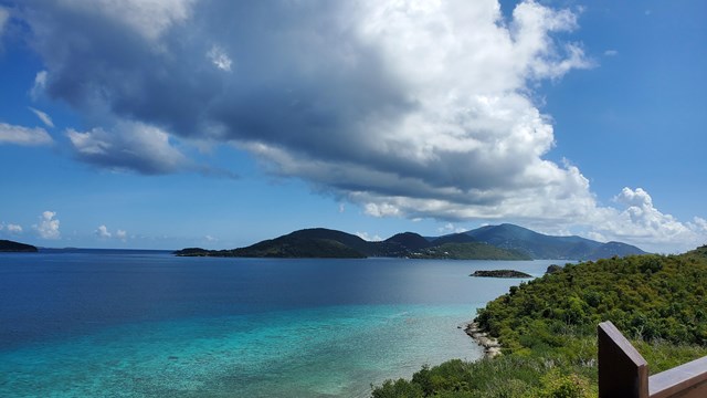 Large white, fluffy clouds soar over a turquoise and blue ocean with shoreline and rail bottom right