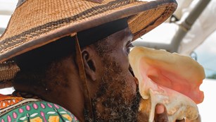 A Black man wearing a bright green shirt and pointed straw hat blowing a white and pink conch shell
