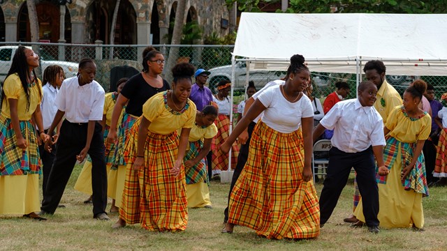 Dancers in traditional yellow and red-checked skirts with men in black pants, white shirts lean in