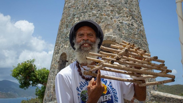 A man demonstrates a traditional fish trap in front of a stone tower.