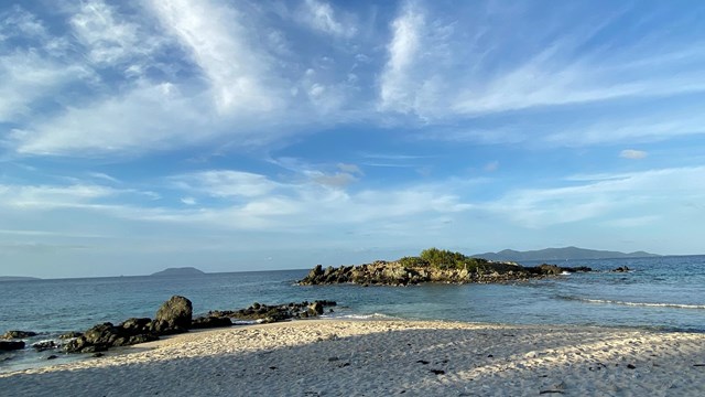 Gentle waves lap the shore of a white sand beach sprinkled with coral and rock.