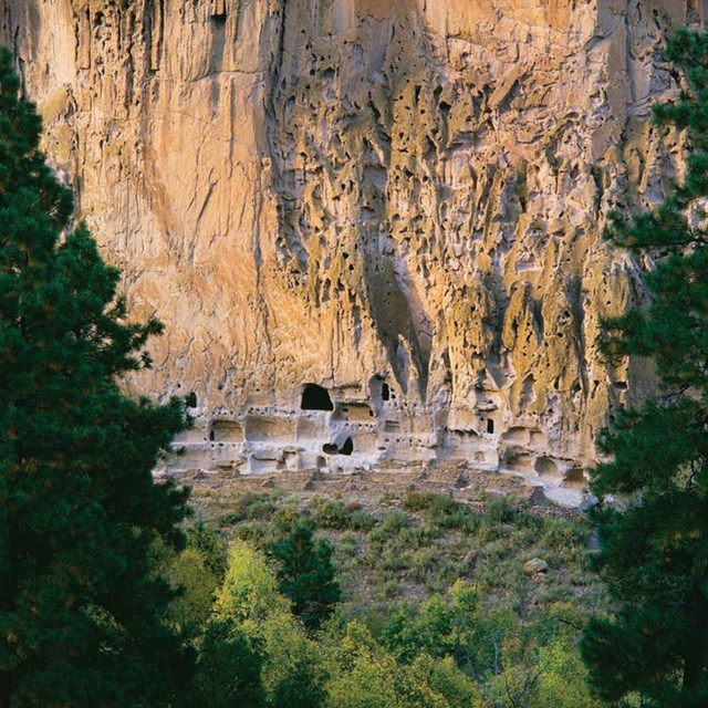Photo of rock cliff with ancient carved out dwellings near the base.