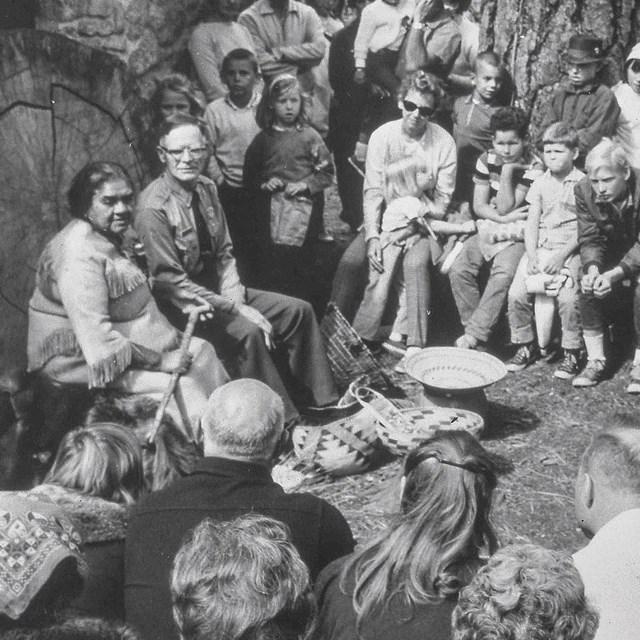 black and white historic photo of a group of people listening to a park program outdoors