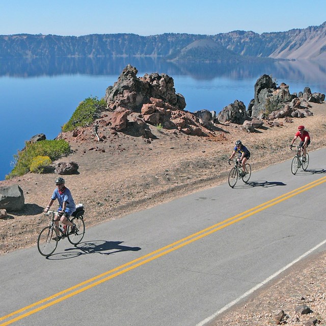 photo of 3 cyclists riding along a scenic highway with crater lake in the distance