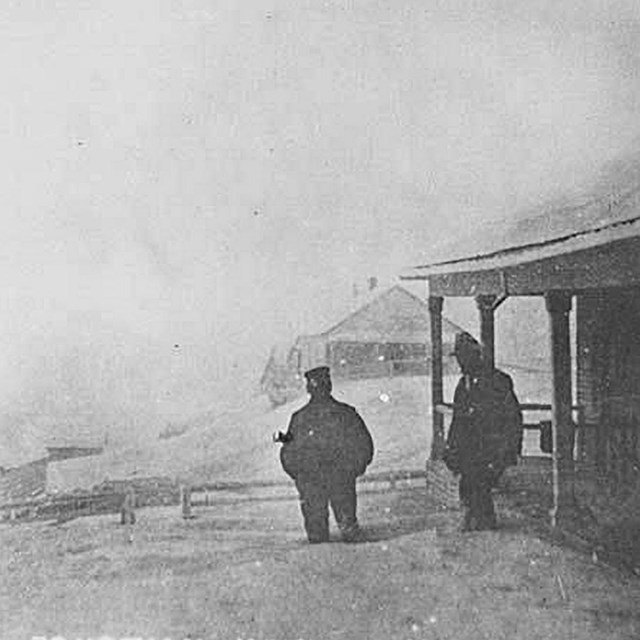 historic black and white photo of two men standing outside a building with deep ashfall deposits all