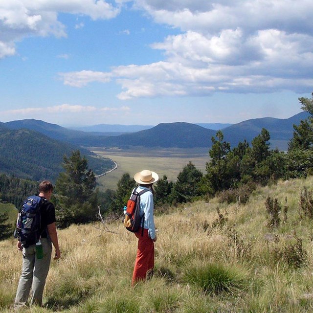photo of two people hiking on a grassy hill slope with mountains and valley in the distance