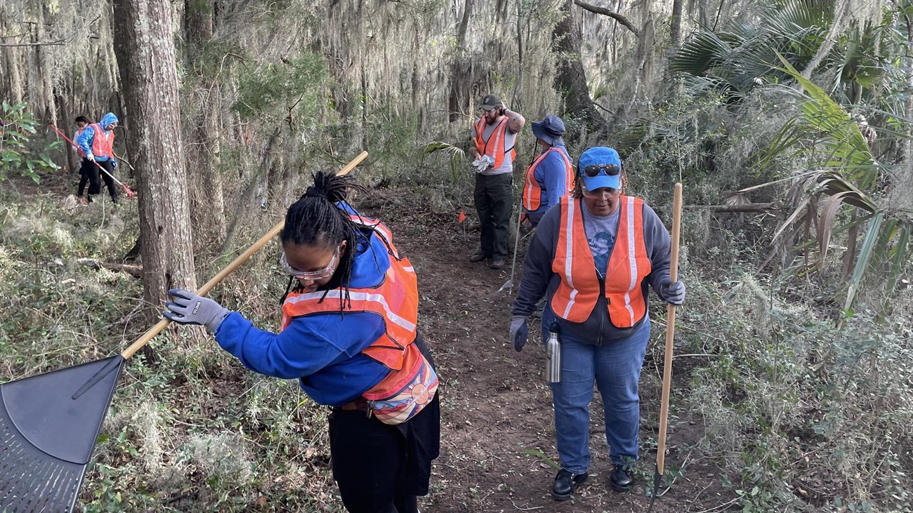 volunteers stand in a forest using hand tools, rakes and shovels to clear a dirt trail
