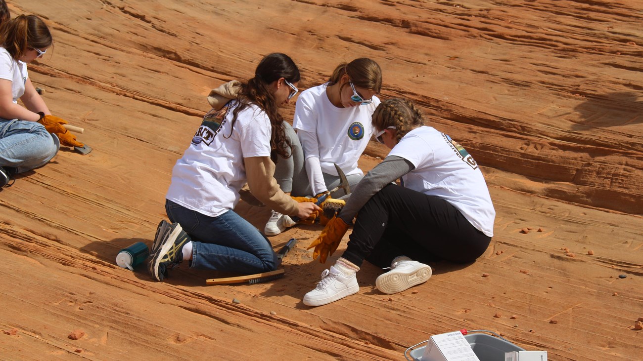 Three teenage volunteers sit in a circle using hand tools to remove graffiti at Glen Canyon National