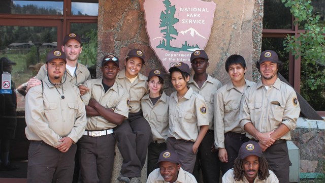 A group of young volunteers in uniform pose for a photo in front of a NPS arrowhead on a building.