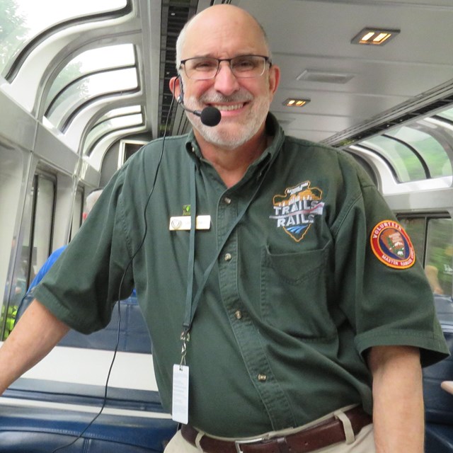 A volunteer guide wearing a headset smiles while standing inside a train.