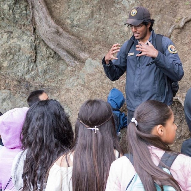 Two adults stand in front of a group of students outdoors.