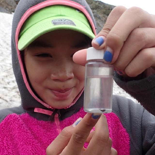 A girl holds a vial of clear liquid on a rocky slope with a patch of snow.