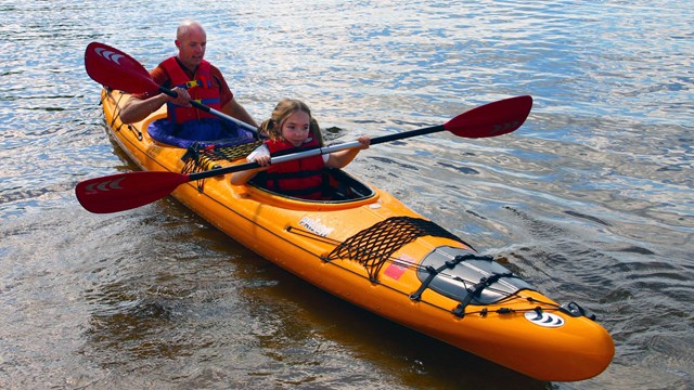 A bright yellow kayak floats near the shore, paddled by a young girl and her father.