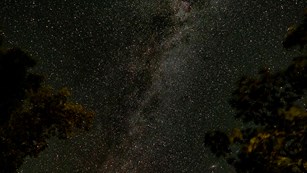 A bright cluster of stars streaks from one horizon to the other across a dark sky.