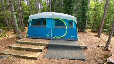 Three wooden stairs lead to a tent pad with a large blue and green tent surrounded by trees.