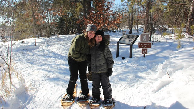 A woman holds her arm over the shoulder of a child.  Both wear snowshoes in front of a snowy trail. 