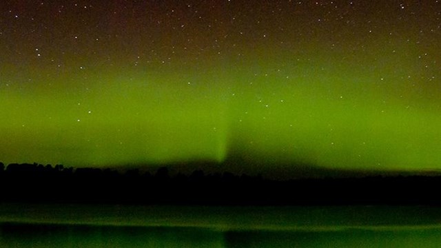 A bright green curtain of light shines near a tree-lined horizon above a scenic lake.