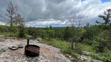 A fire ring sits on a granite ridge surrounded by trees and distant forest.