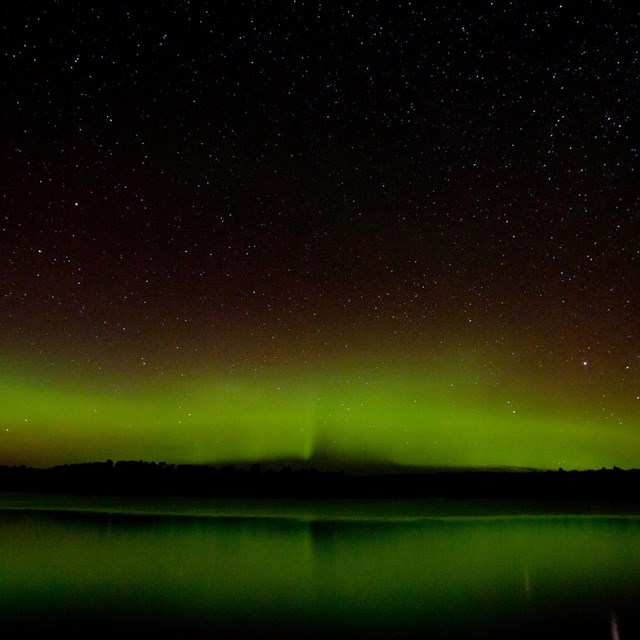 A curtain of green and red light shines against a dark sky, reflected in a large, calm lake.