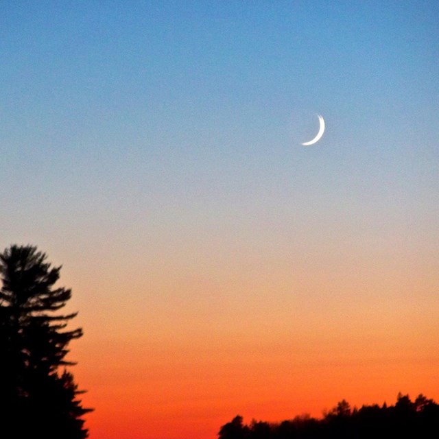A crescent moon shines above a scenic lake, with tree-lined islands silhouetted against a sunset.