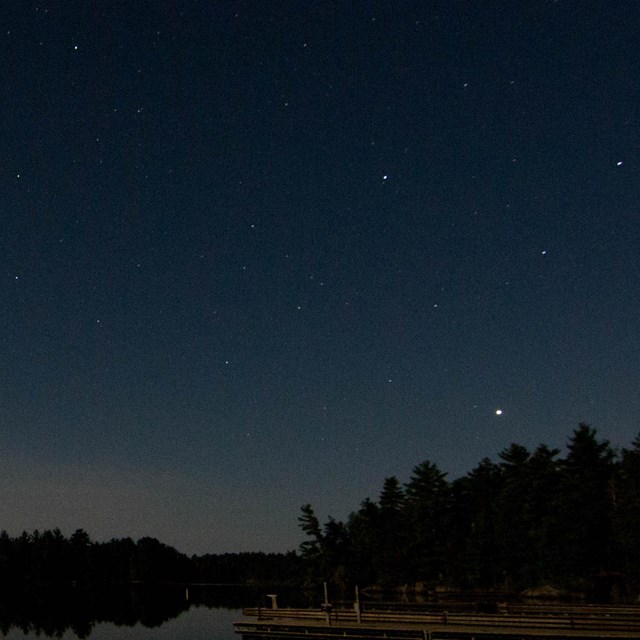 Stars and planets are reflected in the waters of a calm lake with silhouettes of trees on the shore 