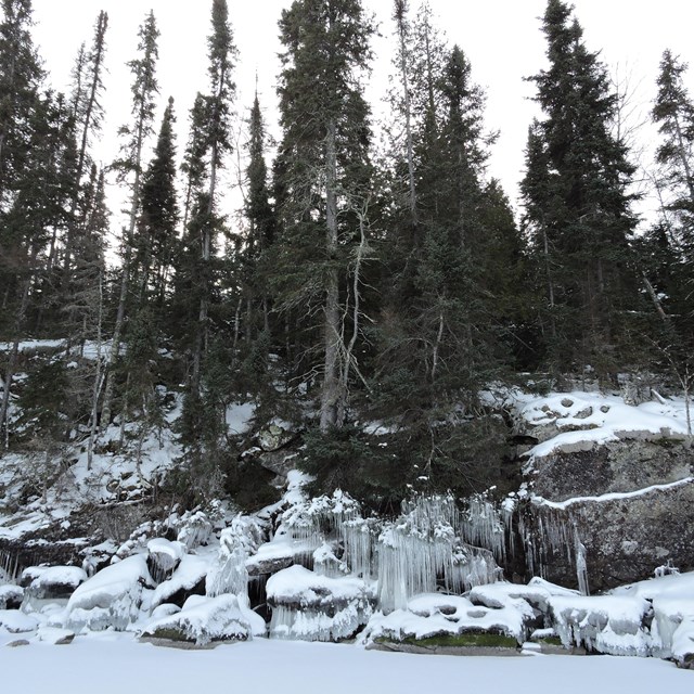 Ice hangs off of rocks on a shoreline of a frozen lake.