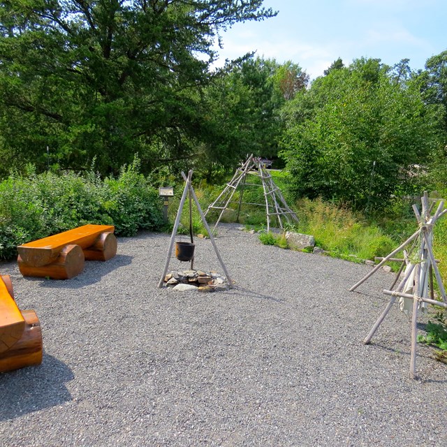 Wooden shelter frame, wooden herb drying rack, kettle hanging from wooden tripod frame over fire pit