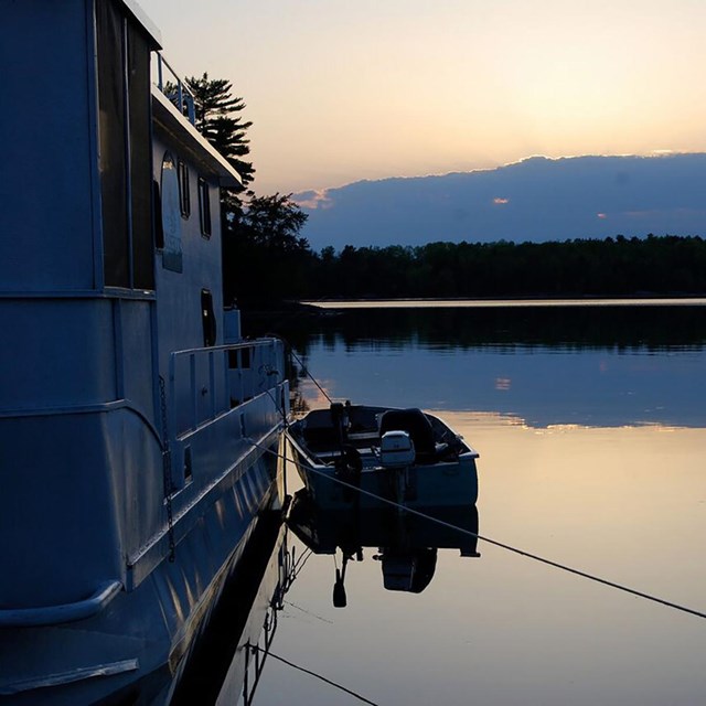 A houseboat and small fishing boat are moored side by side on the shore of a scenic lake at dusk. 