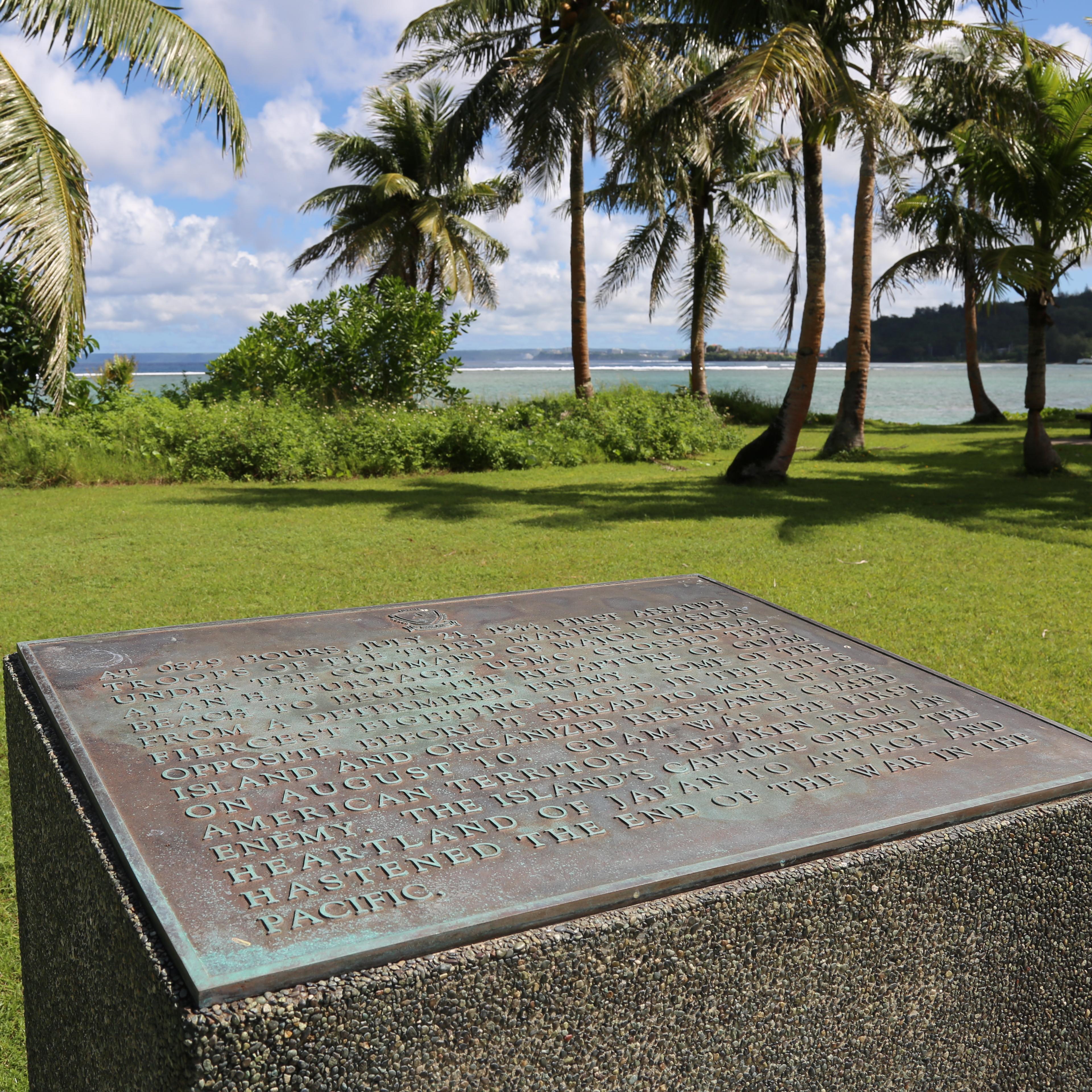 Bronze plaque set on a concrete cube. In the background are palm trees and the ocean.