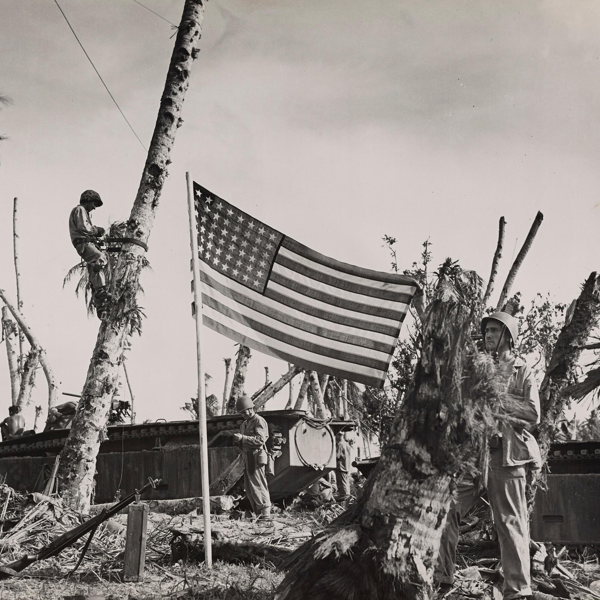 Black and white photo of a solider standing next to a US flag in front of a tank