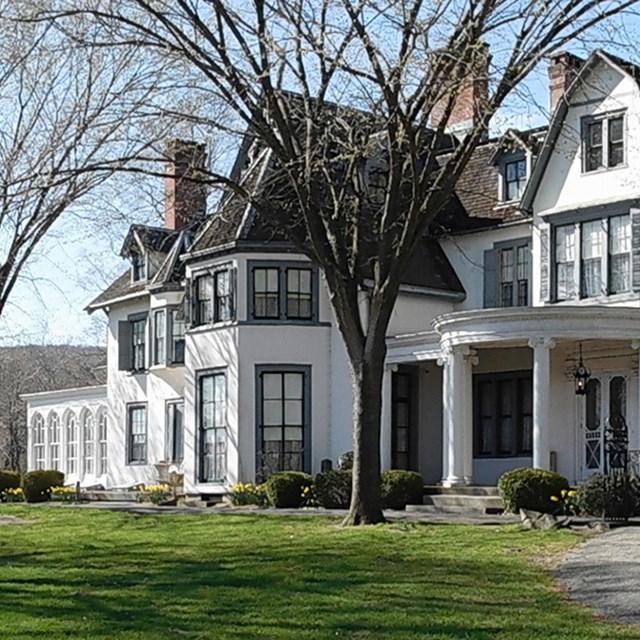 Large white historic mansion with a wraparound front porch, trees in front yard
