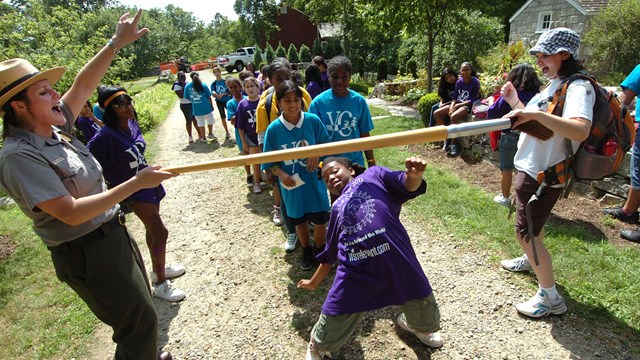 A group of children do the Limbo under a giant paint brush held by park rangers.