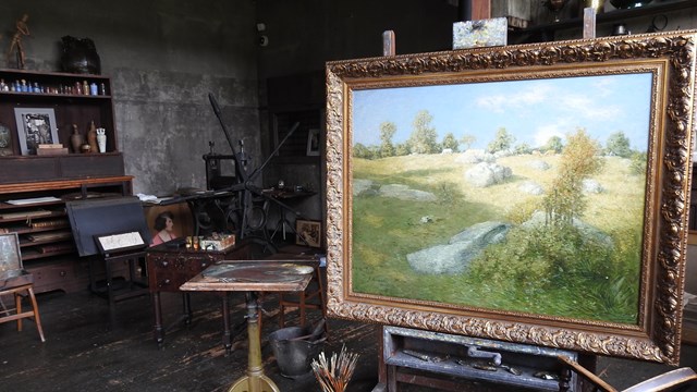 A large painting of a green landscape in a small room filled with easels, chairs, and art supplies.