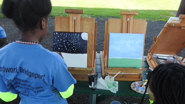 Two young painters sit at easels with partially completed paintings. 