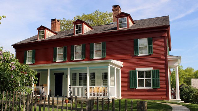 A three-story, red historic home with green shutters and a white porch, among bushes and trees 