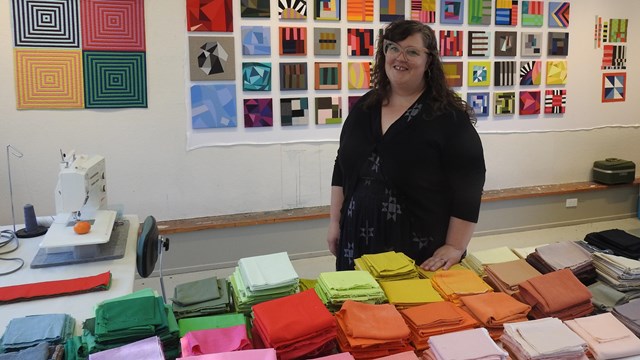 An artist stands behind a table filled with blocks of colorful fabric inside a modern art studio