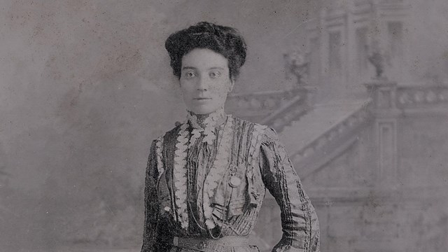 A black and white full length portrait image of a woman standing in a long dress.