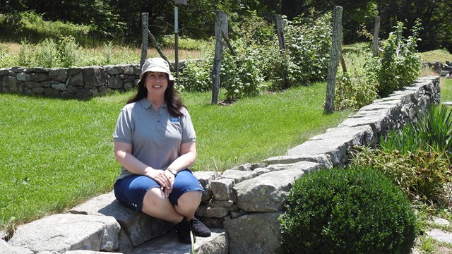 A woman sits on stone steps in a meadow with stone walls surrounding it.
