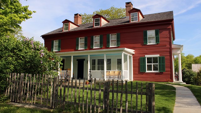 A red house with white trim and green shutters with a wooden fence in the front.